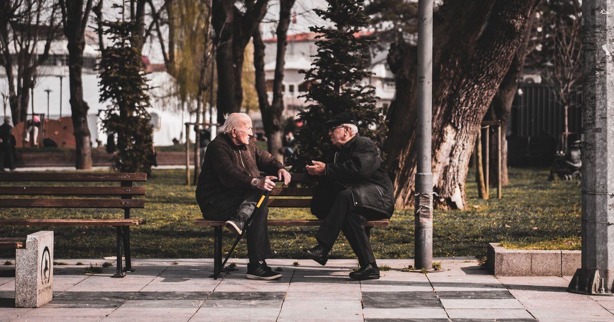 Two people sitting on a park bench, talking outdoors in the afternoon sun