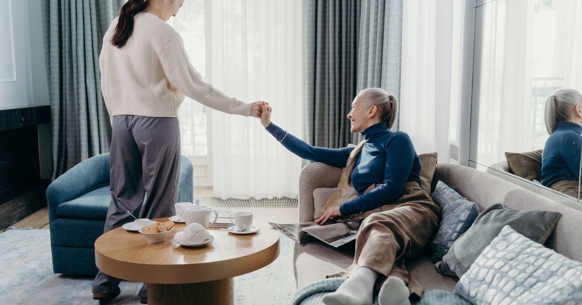 Adult daughter holding hands with her elderly mother in a bright living room